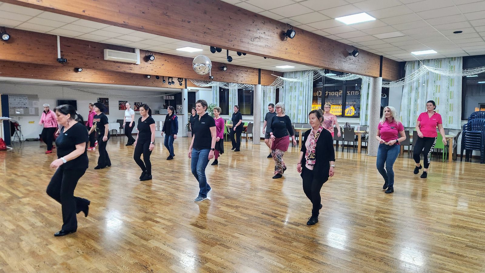 Line-Dance-Training im großen Saal: rund zwanzig Tänzerinnen und Tänzer in Reihen auf dem Parkett, viele in schwarzen oder pinkfarbenen Shirts, darüber Holzbalken, Scheinwerfer und eine Discokugel