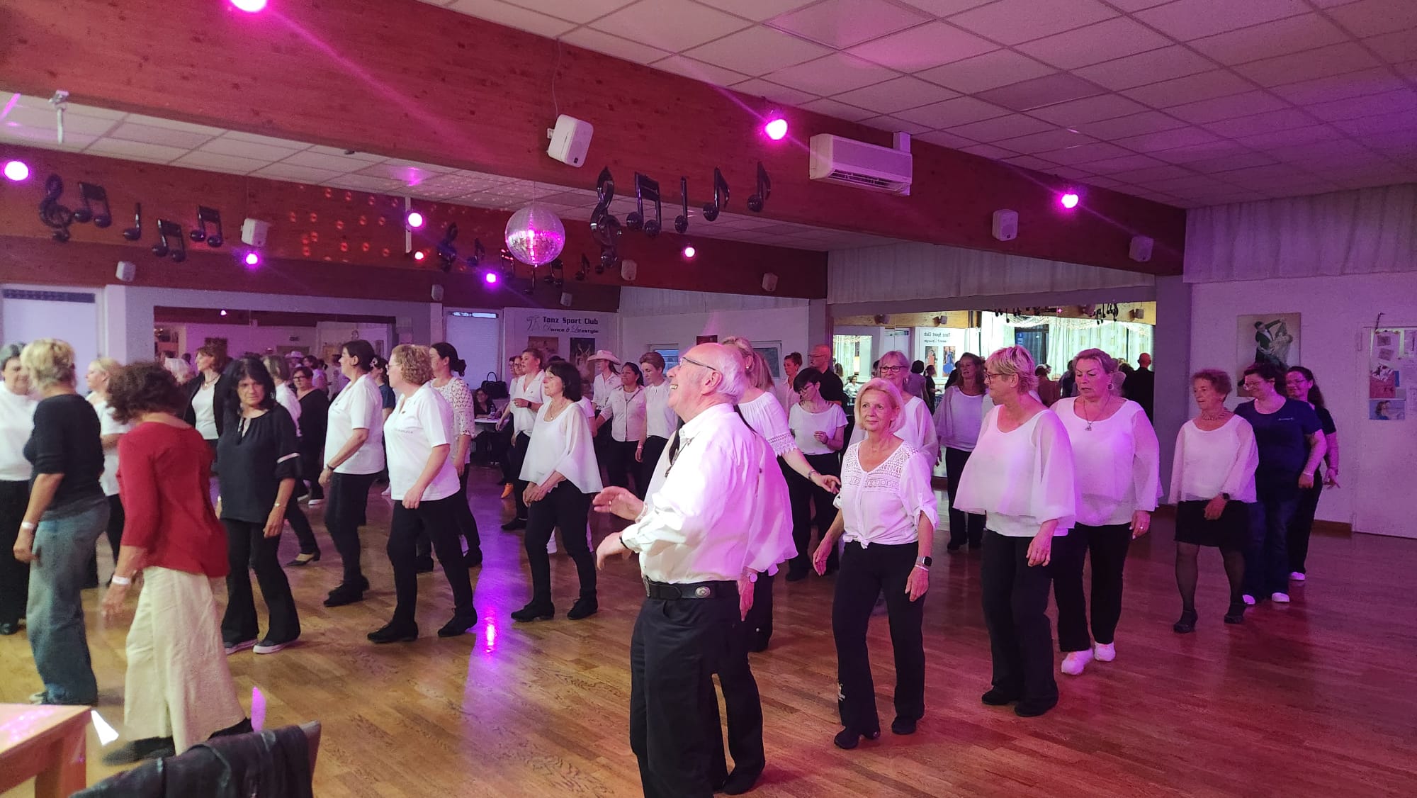 Line-Dance-Gruppe im Saal, viele Tänzerinnen und Tänzer in Reihen auf dem Parkett, darüber Discokugel und Notenschmuck an der Holzdecke